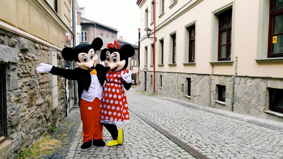 Dwie osoby w kostiumach Myszki Miki i Minnie Mouse stoją na brukowanej ulicy w otoczeniu kamiennych budynków.
