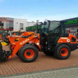 Pomarańczowa ładowarka kołowa Kubota R082 z czarną kabiną i łyżką, stojąca na kostce brukowej przed budynkiem z napisem 'Maszyny Komunalne Budowlane'.