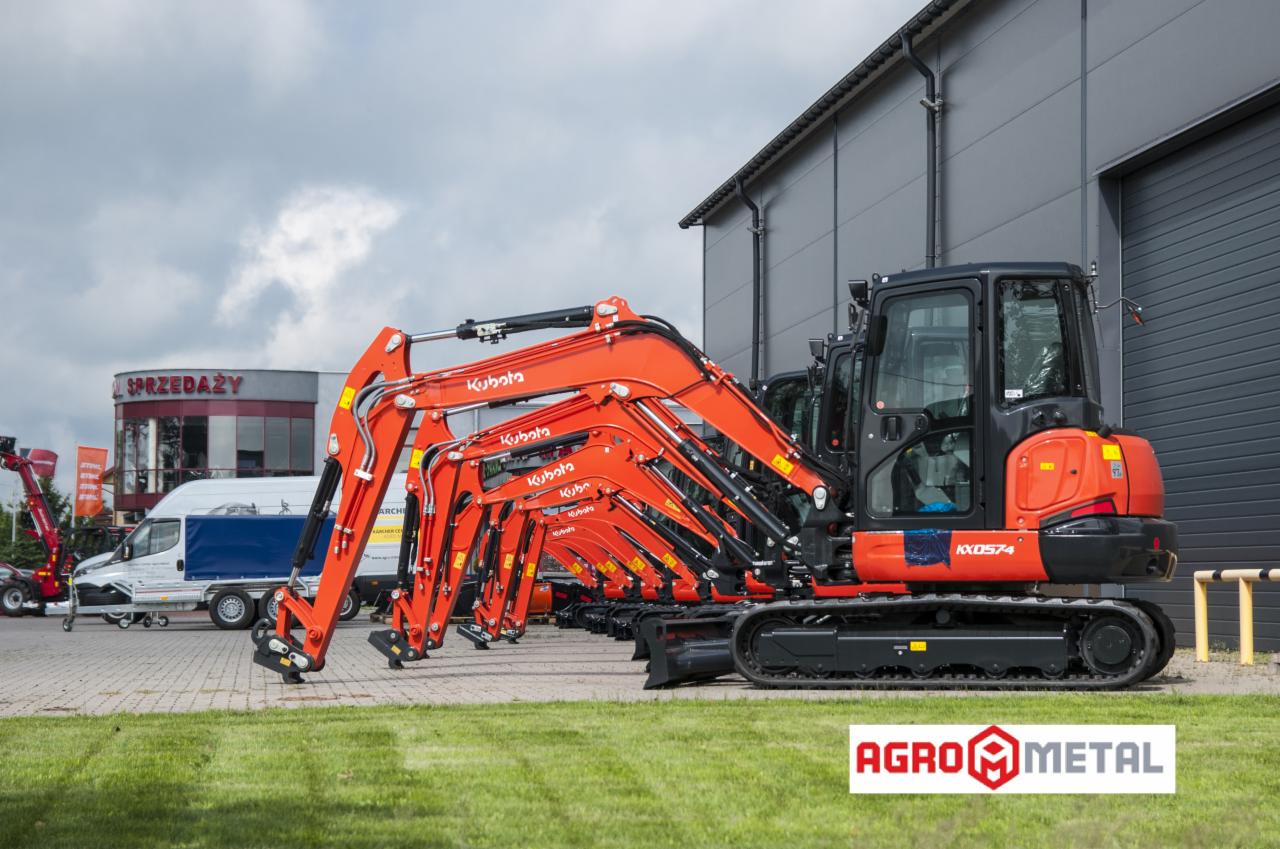 Rząd pomarańczowych, fabrycznie nowych maszyn budowlanych Kubota KX0574 na gąsienicach, ustawionych na trawniku przed szarą halą magazynową i budynkiem sprzedaży.