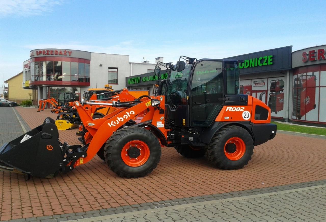 Pomarańczowa ładowarka kołowa Kubota R082 z czarną kabiną i łyżką, stojąca na kostce brukowej przed budynkiem z napisem 'Maszyny Komunalne Budowlane'.