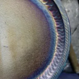 Szczeg&oacute;łowe zbliżenie na okrągły element metalowy z widocznym, precyzyjnym spoiwem po obwodzie, opalizującym odcieniami niebieskiego i fioletu.