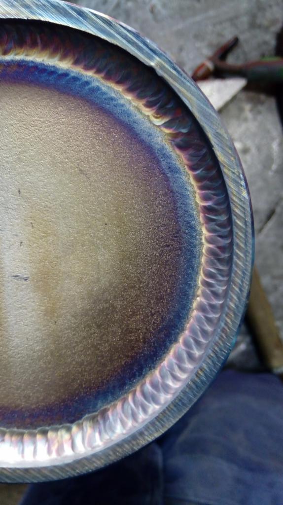 Szczegółowe zbliżenie na okrągły element metalowy z widocznym, precyzyjnym spoiwem po obwodzie, opalizującym odcieniami niebieskiego i fioletu.