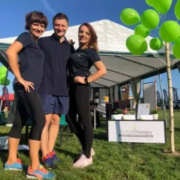 Trzy osoby uśmiechnięte stoją przed stoiskiem ActiveTherapy z zielonymi balonami, na trawie w plenerze.