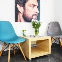 Poczekalnia: Dwa krzesła (turkusowe i szare) przy drewnianym stoliku na kółkach z wazonem białych kwiatów, nad stolikiem portret mężczyzny z brodą.