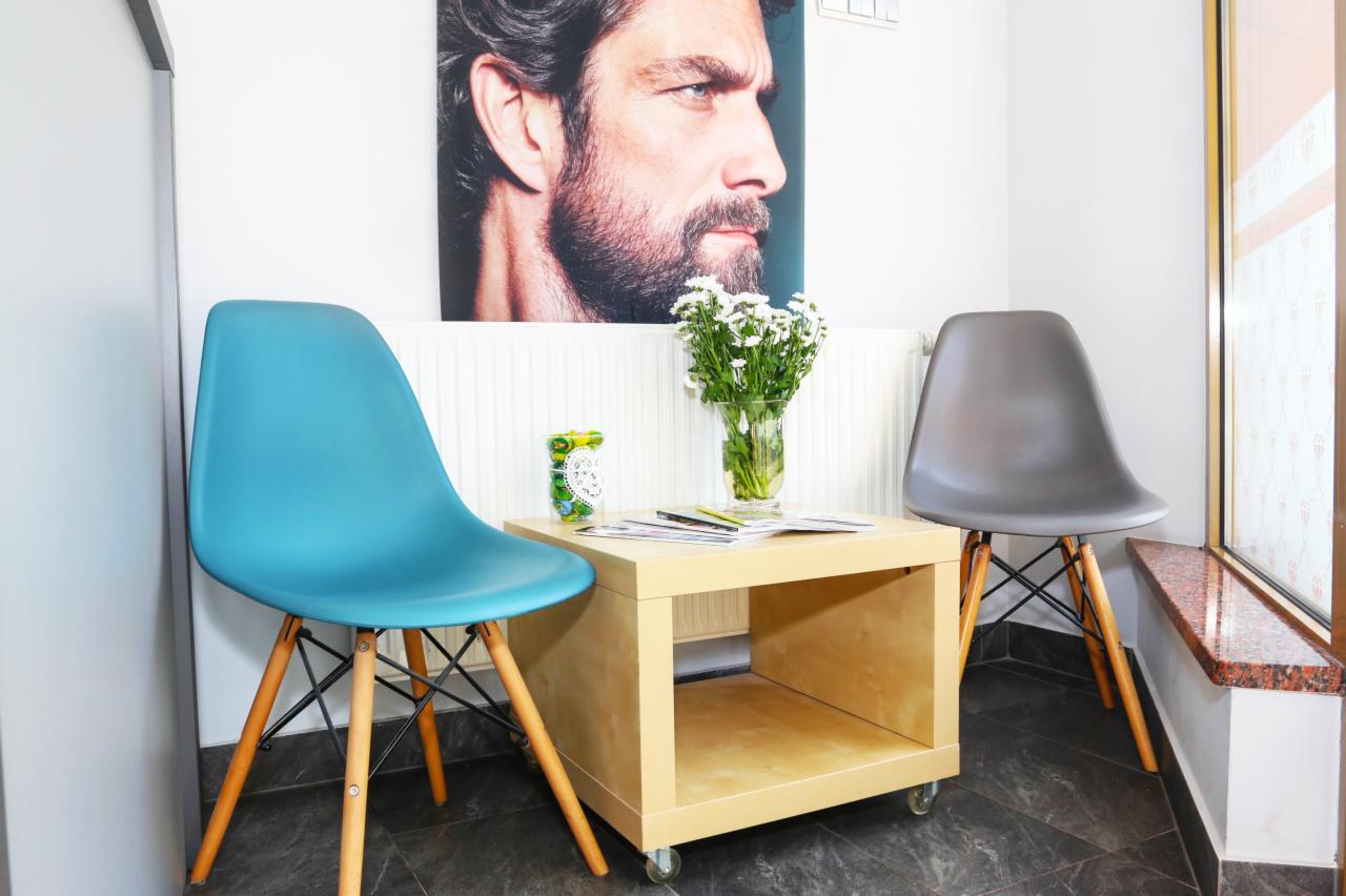 Poczekalnia: Dwa krzesła (turkusowe i szare) przy drewnianym stoliku na kółkach z wazonem białych kwiatów, nad stolikiem portret mężczyzny z brodą.