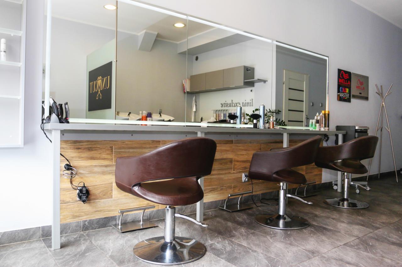 Wnętrze salonu z trzema brązowymi fotelami fryzjerskimi na metalowych podstawach, duże lustro z widocznymi akcesoriami i logo salonu, ściany w odcieniach szarości, płytki podłogowe imitujące kamień.