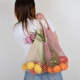 Kobieta z tyłu, niosąca dwie siatki na zakupy: beżową z kwiatami i różową z owocami (pomarańcze, cytryny, pomidory).