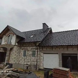 Surowy stan domu z białego pustaka z ciemną dachówką, widoczne rusztowania i stos dachówek, pochmurne niebo w tle.