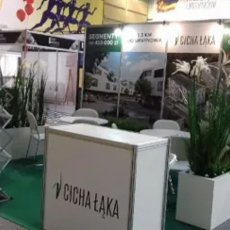 Stoisko targowe dewelopera Cicha Łąka z wizualizacjami segmentów i mieszkań, donicami z trawami i ulotkami informacyjnymi.