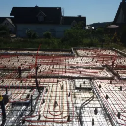 Instalacja ogrzewania podłogowego z rurkami w kolorze czerwonym, ułożonymi w pętle na folii izolacyjnej i siatce zbrojeniowej, widok z góry, w tle zabudowa jednorodzinna.