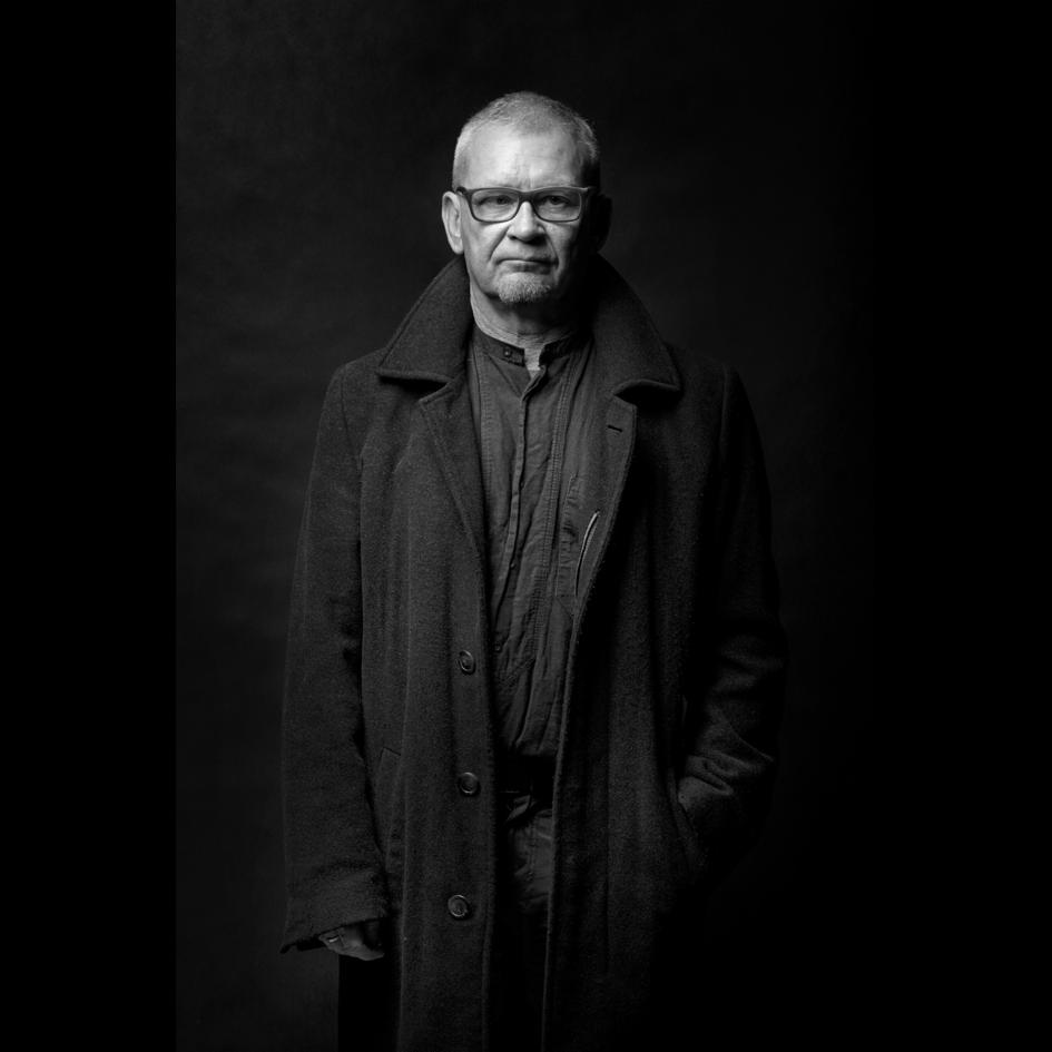 Czarno-biały portret mężczyzny w okularach i płaszczu na ciemnym tle, oświetlony z góry, z rękami w kieszeniach. Elegancki i profesjonalny wizerunek.