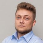 Portret młodego mężczyzny w błękitnej koszuli z rozpiętym górnym guzikiem, z krótkimi włosami i lekkim zarostem na jednolitym, jasnoszarym tle.