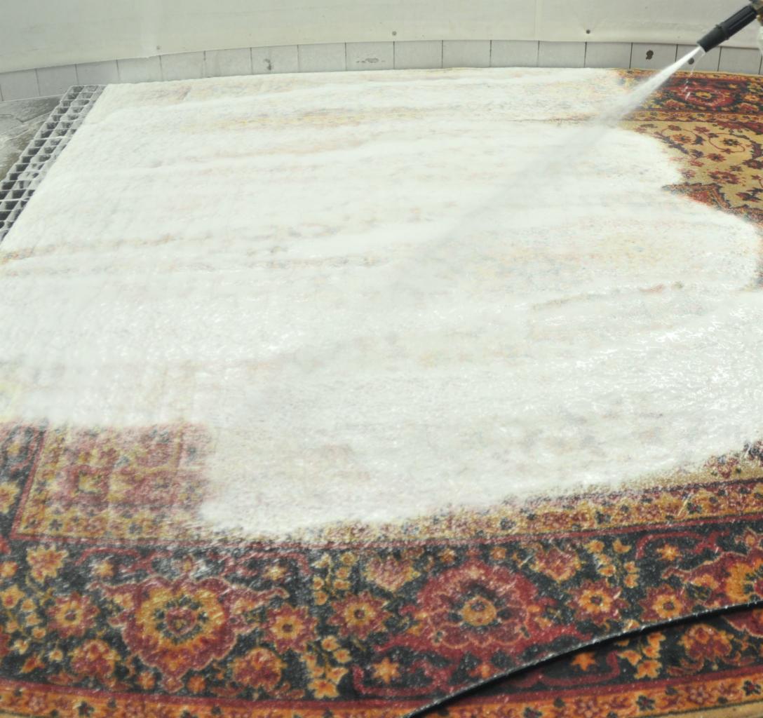 Wzorzysty dywan pokryty pianą podczas czyszczenia pod ciśnieniem, widoczna dysza spryskująca i fragment kratki ściekowej.