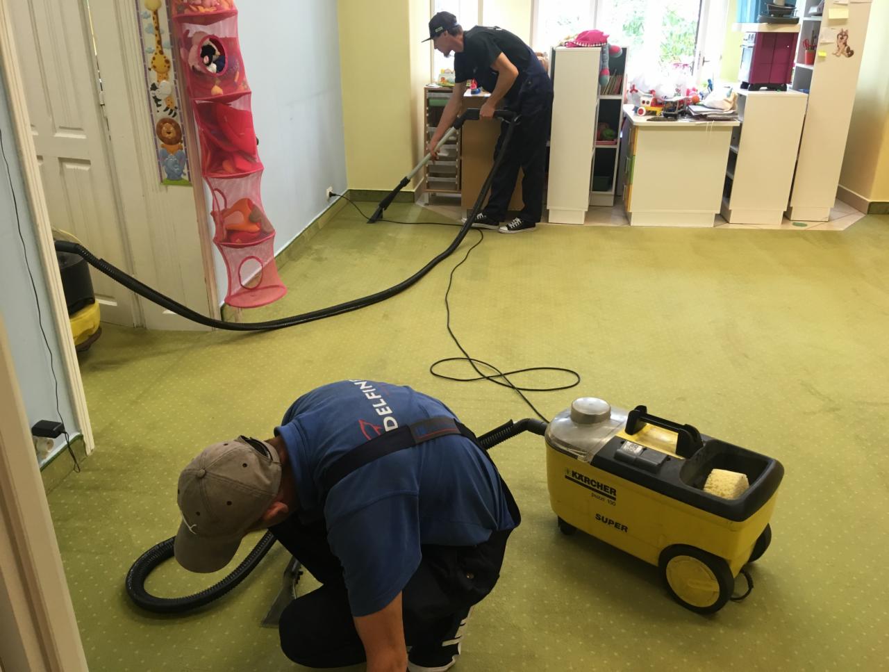 Dwóch mężczyzn w trakcie czyszczenia zielonej wykładziny w pokoju dziecięcym za pomocą odkurzacza piorącego Karcher Puzzi 100 Super; widoczny kosz na zabawki i białe szafki.