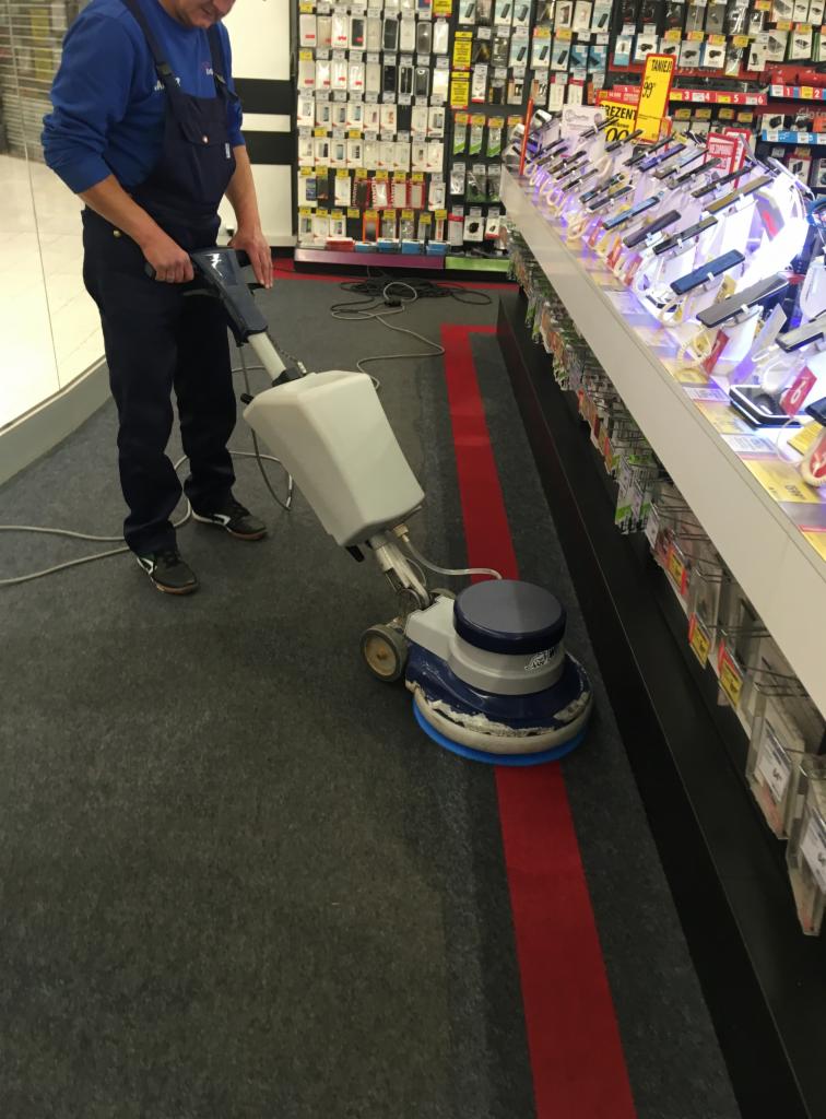 Pracownik w niebieskim kombinezonie obsługuje maszynę czyszczącą szary dywan z czerwonym pasem w sklepie z elektroniką, w tle półki z akcesoriami telefonicznymi.