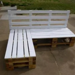 Biała narożna sofa ogrodowa wykonana z palet, ustawiona na betonowym patio, widoczna trawa w tle.