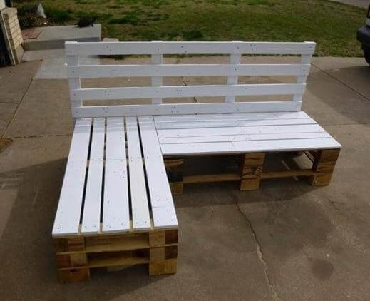 Biała narożna sofa ogrodowa wykonana z palet, ustawiona na betonowym patio, widoczna trawa w tle.