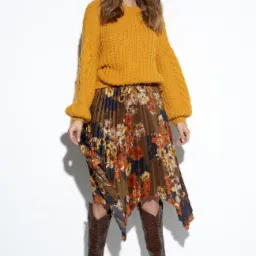 Modelka w musztardowym swetrze z warkoczowym splotem, plisowanej spódnicy w kwiaty i brązowych kozakach ze skóry węża na białym tle.