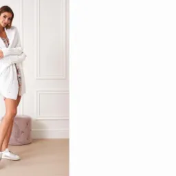 Uśmiechnięta kobieta w białym, dzierganym kardiganie z rękawami 3/4, białych sneakersach i kwiecistej sukience pozuje w jasnym wnętrzu z dekoracyjnymi sztukateriami, obok stolika z orchideą i pufy.