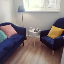 Kącik relaksacyjny z granatową sofą, pastelowymi poduszkami, fotelem i stolikiem z wodą w jasnym gabinecie.