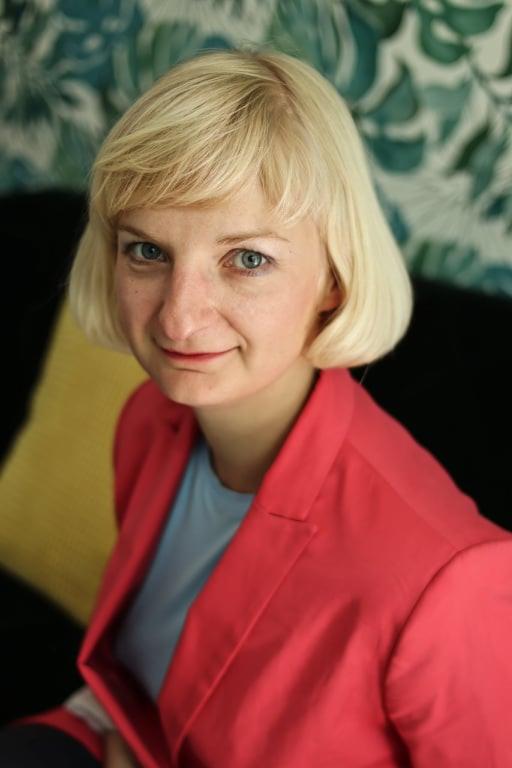 Portret kobiety z blond włosami w koralowej marynarce na tle wzorzystej tapety.