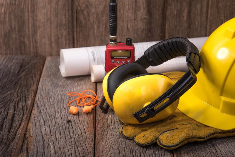 Żółty kask ochronny, słuchawki przeciwhałasowe, pomarańczowe zatyczki do uszu, rękawice robocze, czerwone radio i zwój planów na drewnianym stole.