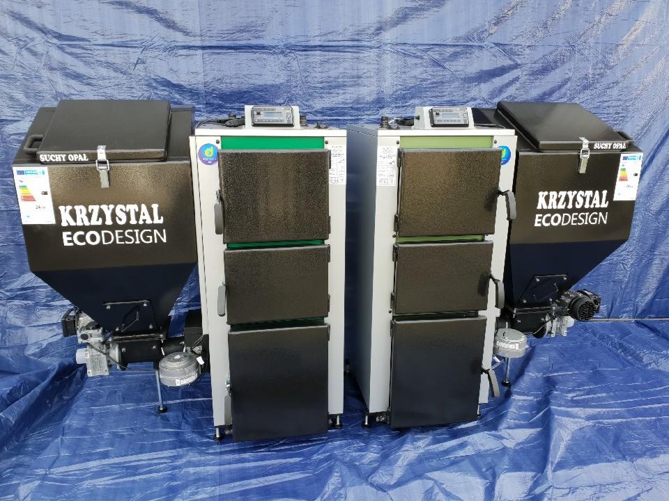Dwa piece na paliwo stałe marki KRZYSTAL ECODESIGN, ustawione obok siebie na niebieskiej plandece, widoczne zbiorniki na paliwo i panele sterowania.