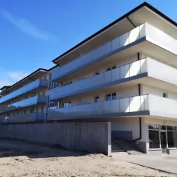 Nowoczesny budynek apartamentowy z balkonami zabezpieczonymi szkłem mlecznym, widok z poziomu gruntu na plac budowy.