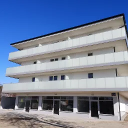 Nowoczesny, trzypiętrowy budynek z białymi ścianami i balkonami zabezpieczonymi mlecznymi taflami szkła, widoczny numer adresowy 18, otoczenie w stanie surowym.