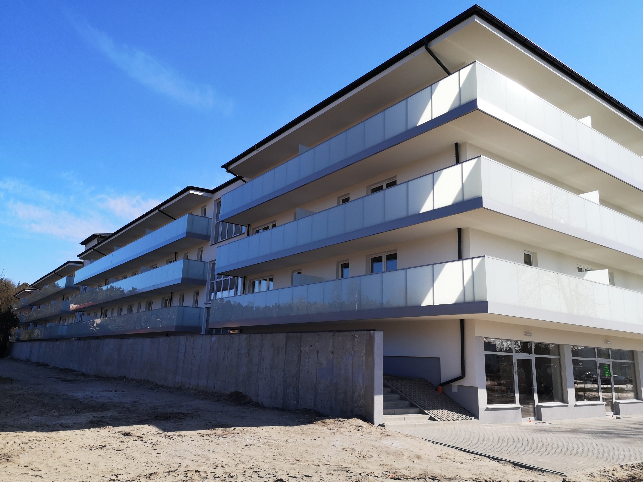 Nowoczesny budynek apartamentowy z balkonami zabezpieczonymi szkłem mlecznym, widok z poziomu gruntu na plac budowy.