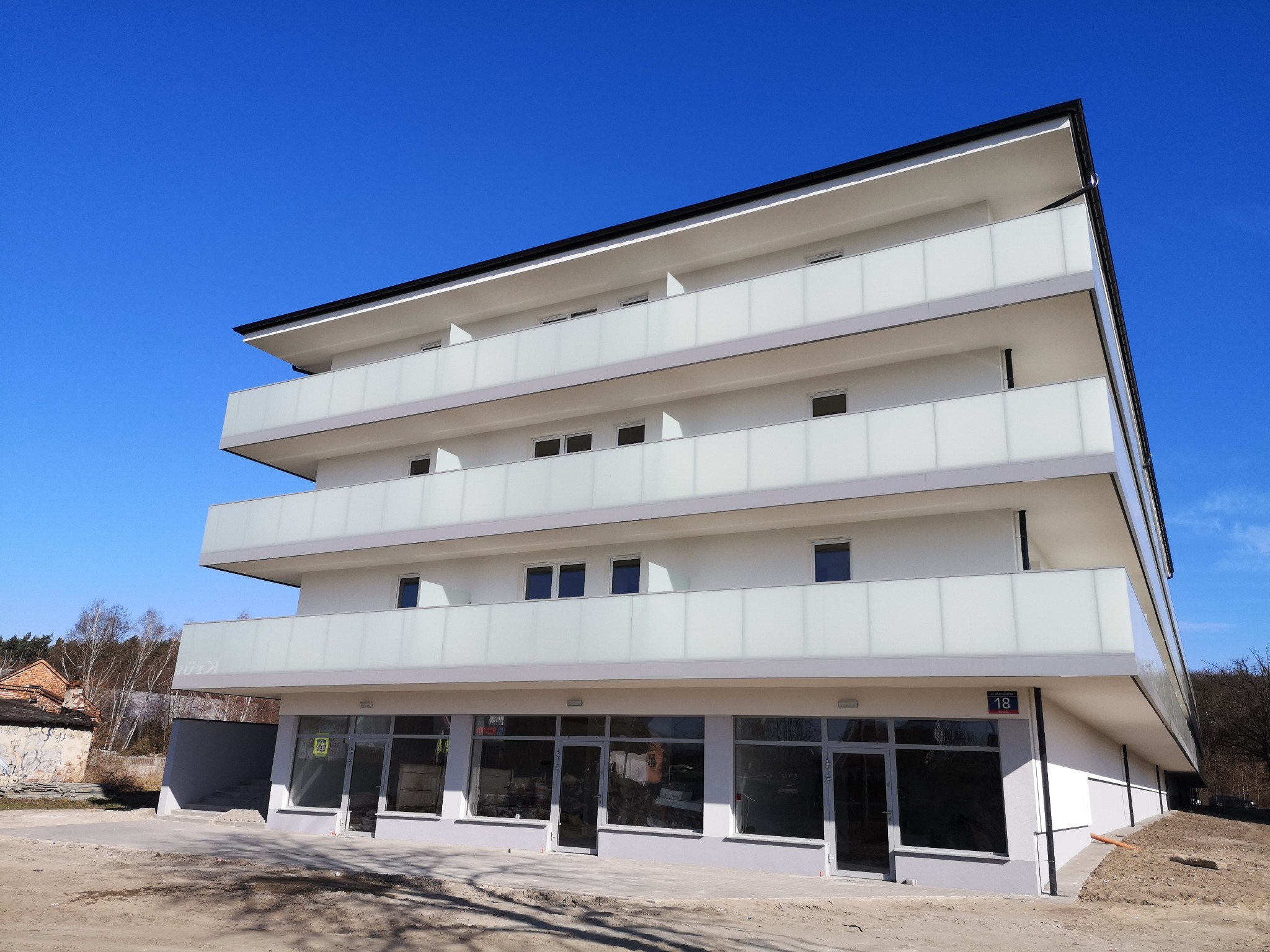 Nowoczesny, trzypiętrowy budynek z białymi ścianami i balkonami zabezpieczonymi mlecznymi taflami szkła, widoczny numer adresowy 18, otoczenie w stanie surowym.