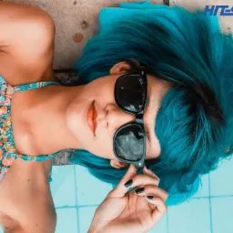 Kobieta z turkusowymi włosami i okularami przeciwsłonecznymi leży obok basenu, w tle logo 'HIT SPORT travel'.