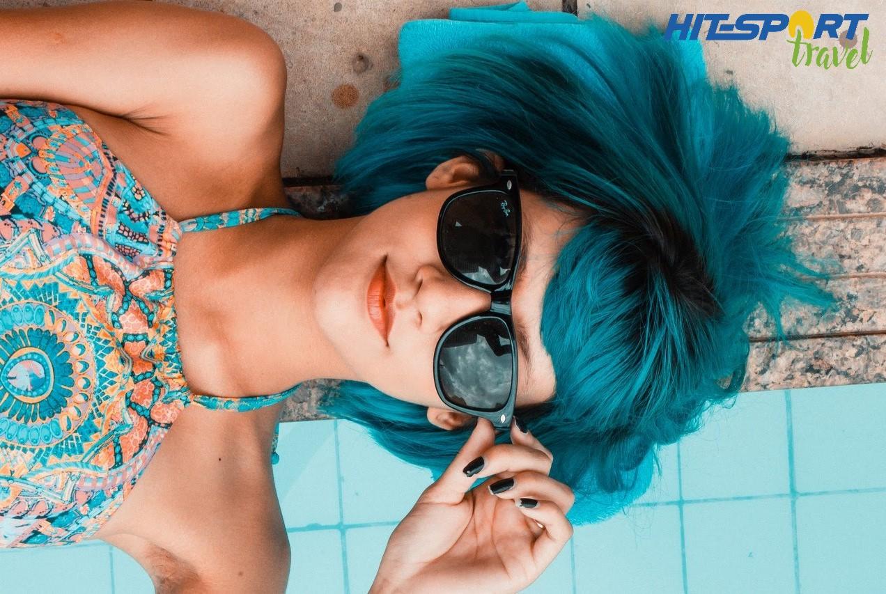 Kobieta z turkusowymi włosami i okularami przeciwsłonecznymi leży obok basenu, w tle logo 'HIT SPORT travel'.