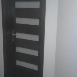 rubin - Ciemne, nowoczesne drzwi wewnętrzne z pięcioma poziomymi, matowymi przeszkleniami, zamontowane w białej ścianie na podłodze imitującej ciemne drewno.