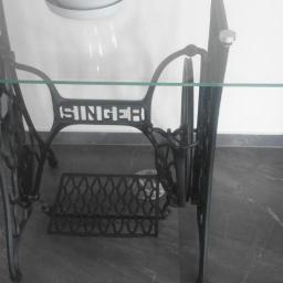 rubin - Stara, żeliwna podstawa maszyny do szycia Singer zaadaptowana jako stolik z blatem szklanym, widoczna na tle białej ściany i doniczki z rośliną.