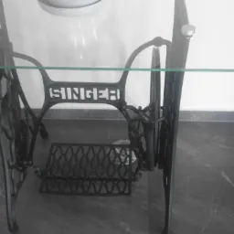 Stara, żeliwna podstawa maszyny do szycia Singer zaadaptowana jako stolik z blatem szklanym, widoczna na tle białej ściany i doniczki z rośliną.