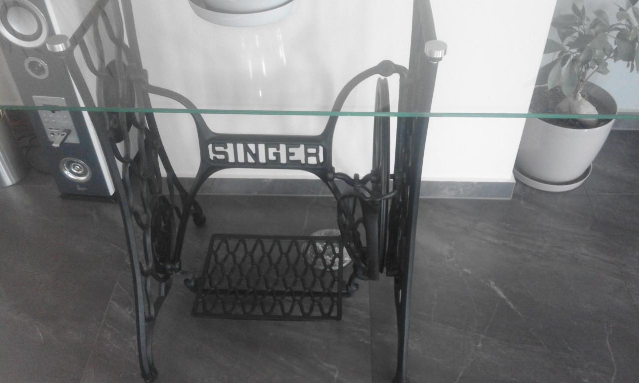 Stara, żeliwna podstawa maszyny do szycia Singer zaadaptowana jako stolik z blatem szklanym, widoczna na tle białej ściany i doniczki z rośliną.