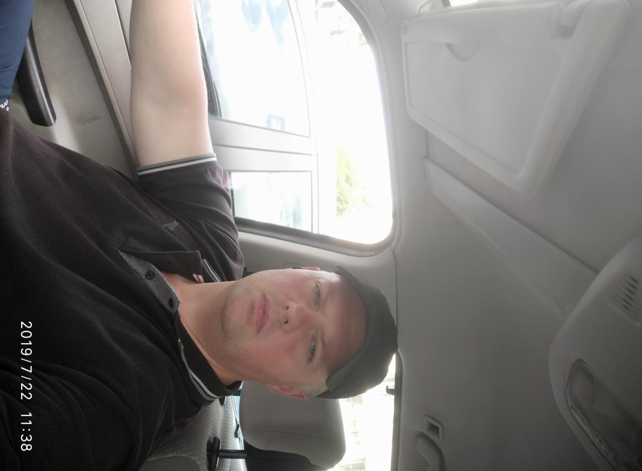 Mężczyzna w czarnej koszulce polo i czapce z daszkiem siedzi w jasnym samochodzie, widoczny przez okno fragment zieleni. Zdjęcie zrobione z perspektywy pasażera.