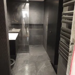 Nowoczesna łazienka z ciemnymi płytkami imitującymi marmur, kabina prysznicowa typu walk-in, wisząca umywalka z szafką oraz grzejnik drabinkowy w kolorze czarnym.