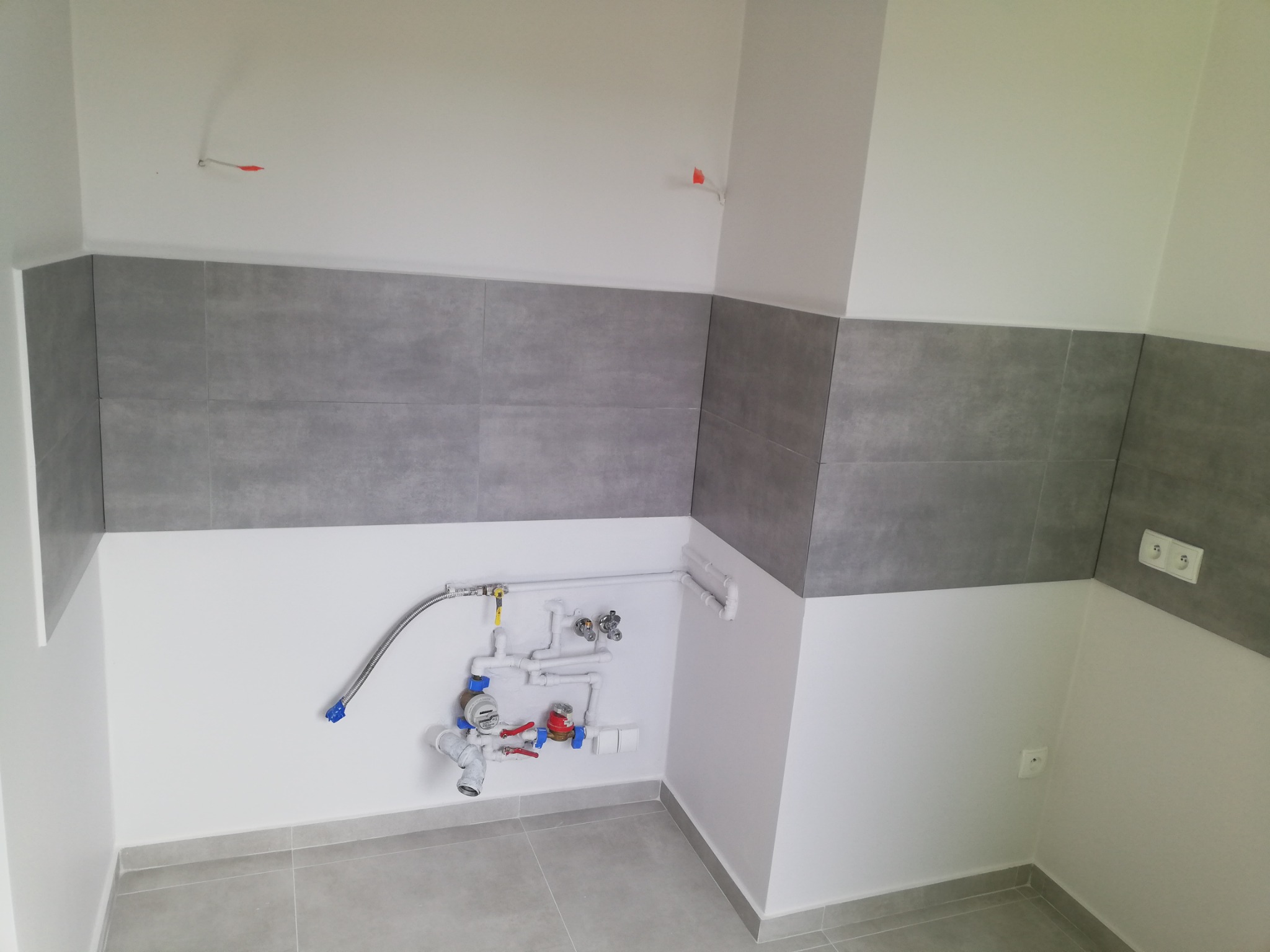 Szare płytki ścienne i podłogowe w trakcie remontu łazienki, widoczne rury i instalacja wodna, przygotowane do podłączenia urządzeń sanitarnych, fragment ściany z gniazdkami.