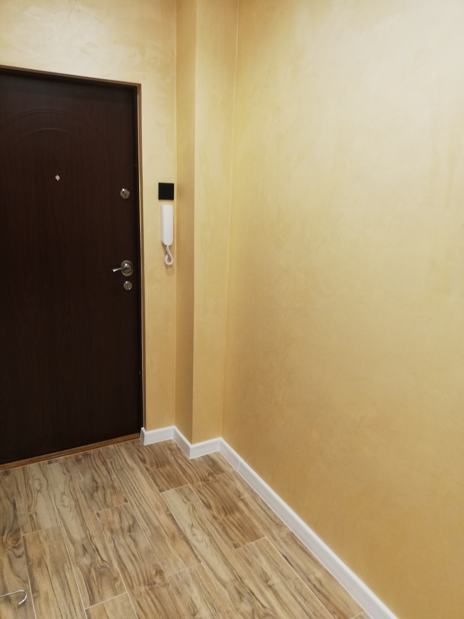 Wnętrze przedpokoju z brązowymi drzwiami wejściowymi, żółtymi ścianami z dekoracyjną fakturą, listwami przypodłogowymi i panelami podłogowymi imitującymi drewno.