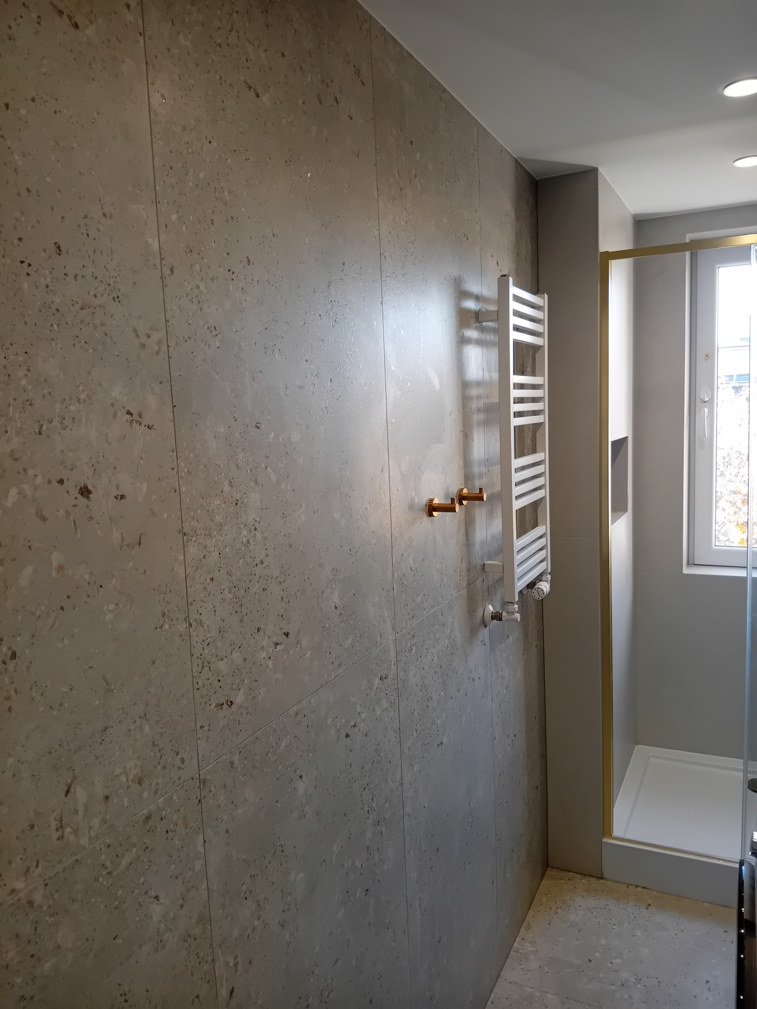 Wykończona łazienka z szarymi płytkami imitującymi lastryko na ścianach i podłodze, biały grzejnik drabinkowy z miedzianymi zaworami, kabina prysznicowa z złotymi profilami.