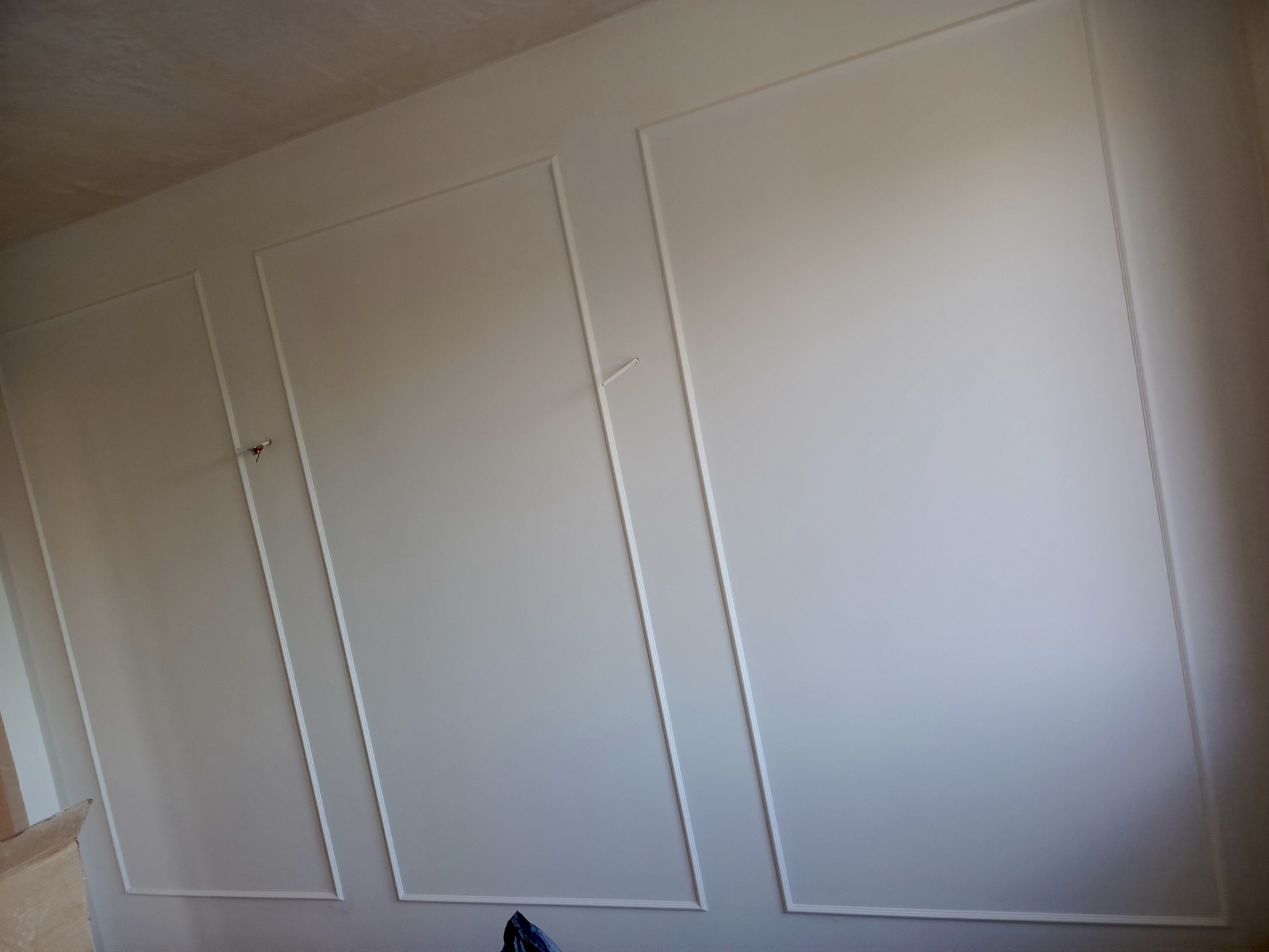 Ściana z zamontowanymi białymi panelami dekoracyjnymi, widoczne niedoskonałości wykończenia i wystające przewody.