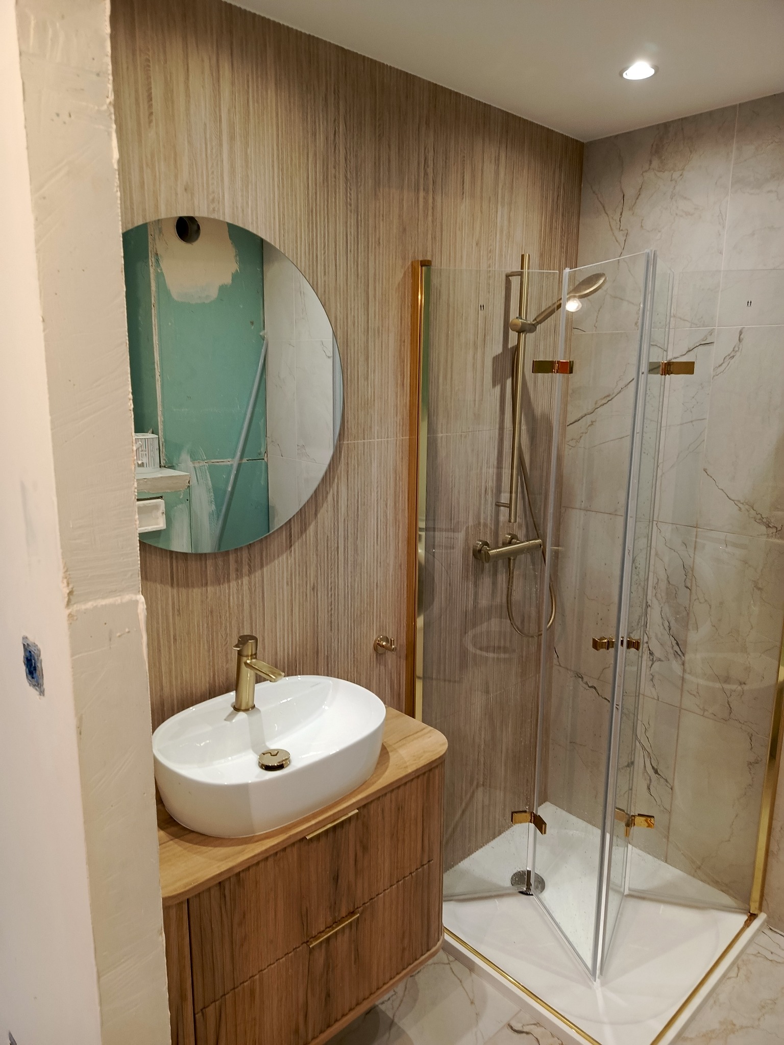 Nowoczesna łazienka w trakcie wykańczania: owalne umywalka na drewnianej szafce z złotą armaturą, kabina prysznicowa z złotymi detalami, okrągłe lustro z widoczną ścianą z płyt karton-gips.