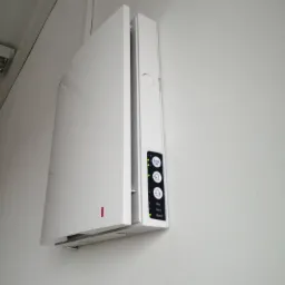 Nowoczesny, biały oczyszczacz powietrza z panelem sterowania LED, zamontowany na białej ścianie. Widoczne diody sygnalizujące tryb pracy i stan filtra.