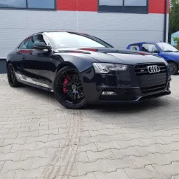 Błyszczący, czarny samochód Audi S5 Coupe z czerwonymi zaciskami hamulcowymi na tle szarego budynku z czerwonymi akcentami i kostki brukowej.
