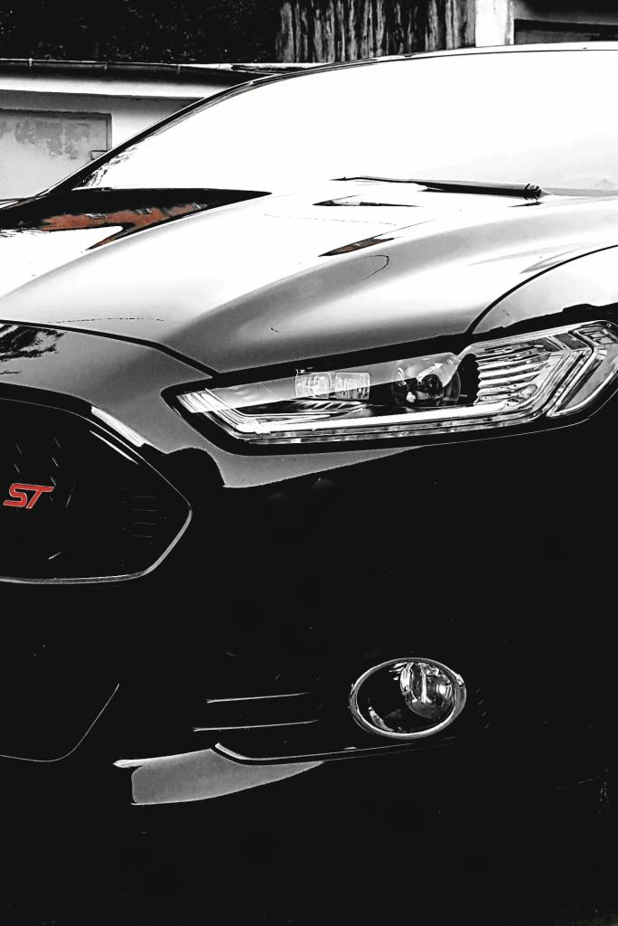 Częściowy widok przodu czarnego samochodu Ford Focus ST z charakterystycznym czerwonym logo ST na grillu, uchwycony w czerni i bieli z elementami koloru, skupiający się na detalu reflektora i lampy...