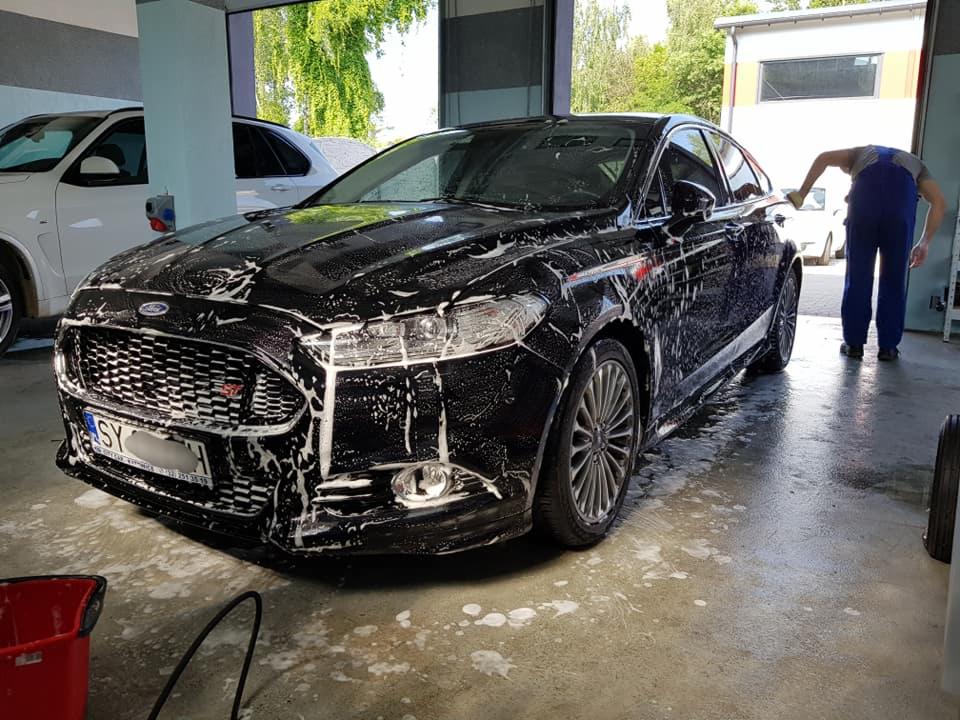 Czarny Ford Mondeo pokryty pianą podczas mycia w myjni; pracownik w niebieskim kombinezonie pochyla się nad samochodem.