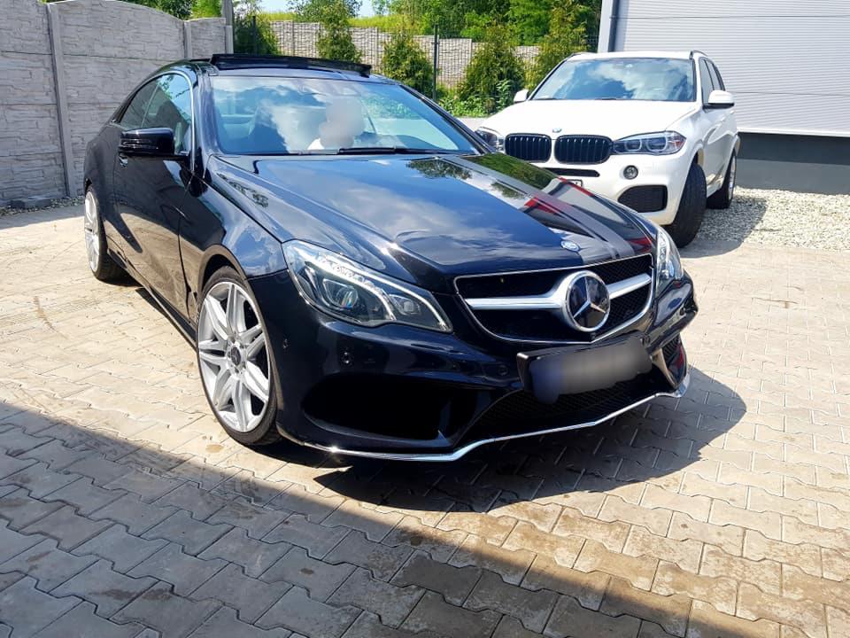 Błyszczący, czarny Mercedes E-klasa Coupe z otwartym szyberdachem zaparkowany na kostce brukowej, z białym BMW X5 w tle.