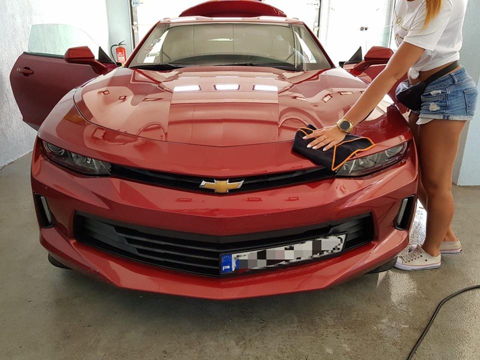 Czerwony Chevrolet Camaro z otwartymi drzwiami, wycierany czarną ściereczką z pomarańczowym obszyciem przez kobietę w dżinsowych szortach w myjni samochodowej.